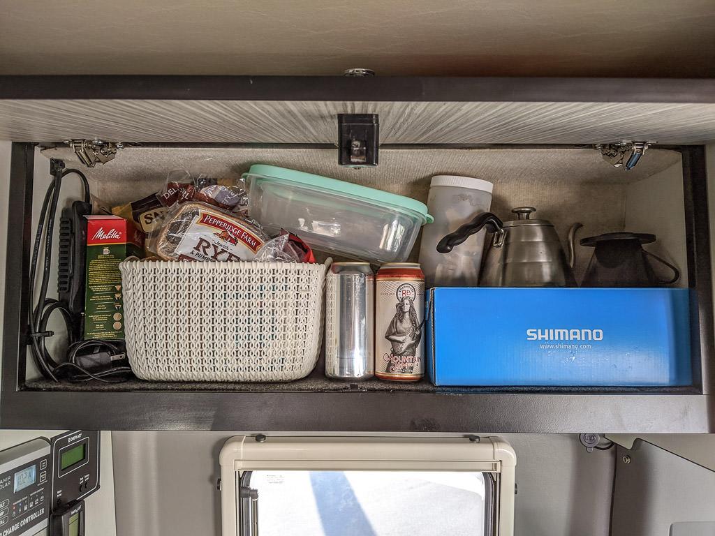 Cabinet above bench seat with shoes box full of spices, coffee making supplies, bread and snacks