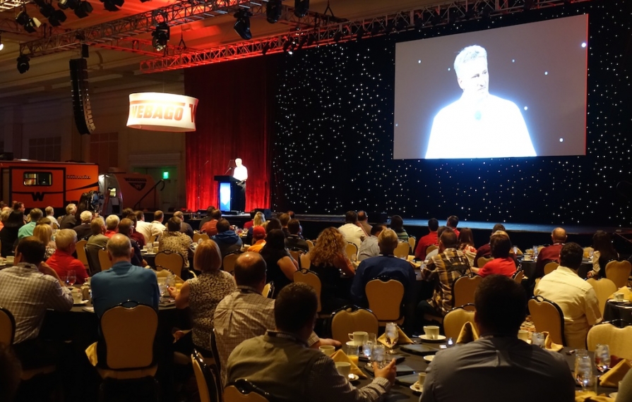 Crowd at tables as speaker talks on stage at Dealer Days.