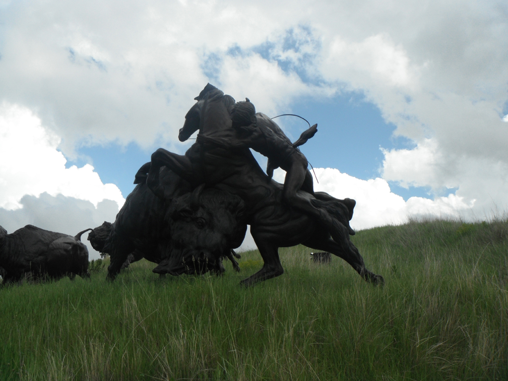 Statue of indians and bison in tall grassy plain.