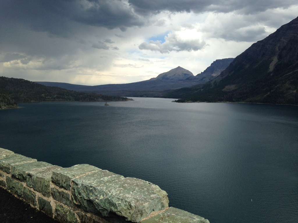 Stone bridge alongside waterway with mountains on the other side and a cloudy sky above.