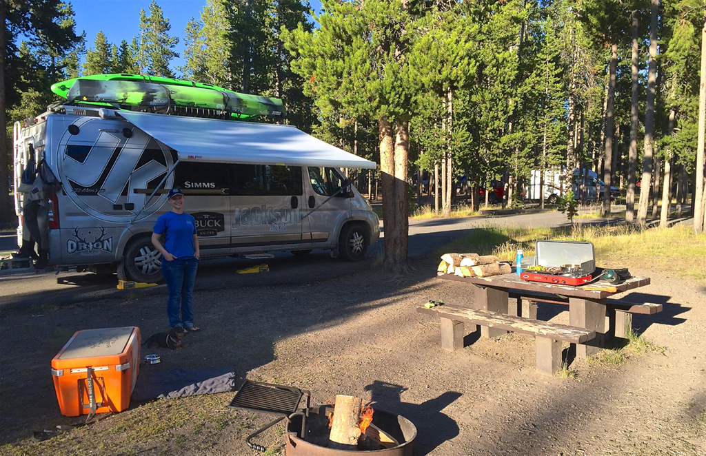 Winnebago Travato parked in a campsite with awning out and a picnic table and campfire next to it.