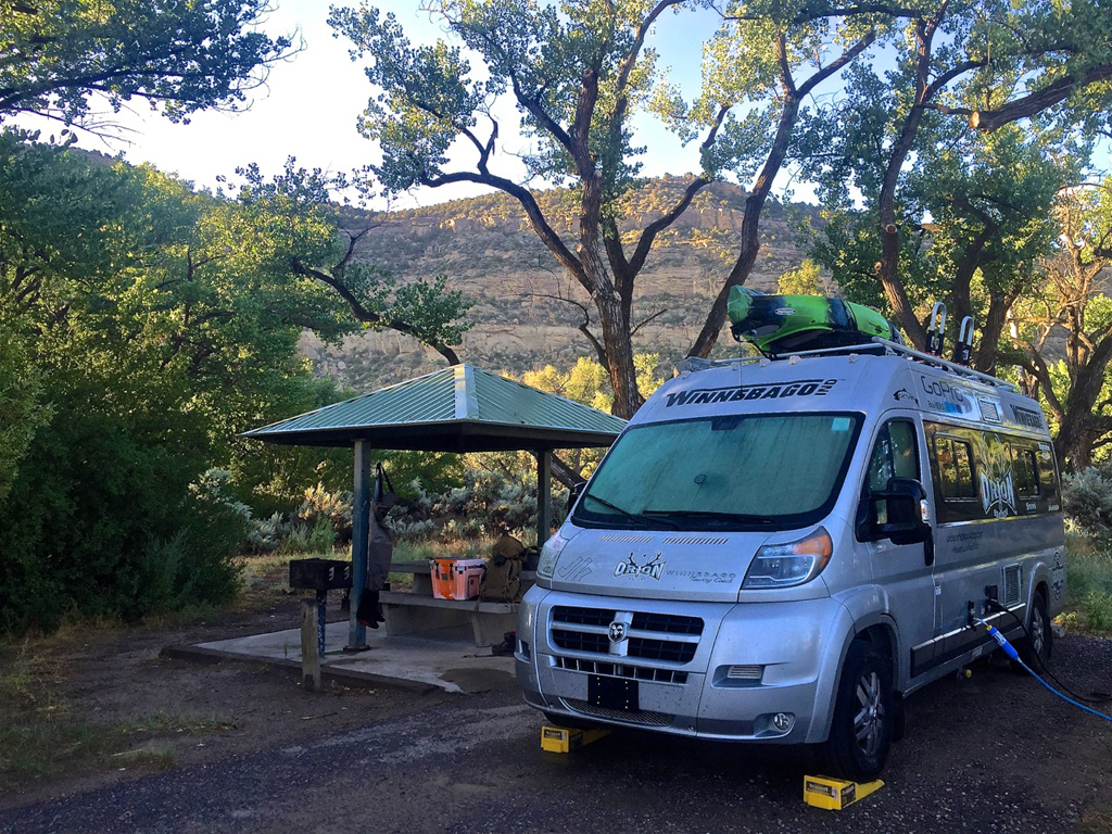 Winnebago Travato parked in campsite with trees surrounding.