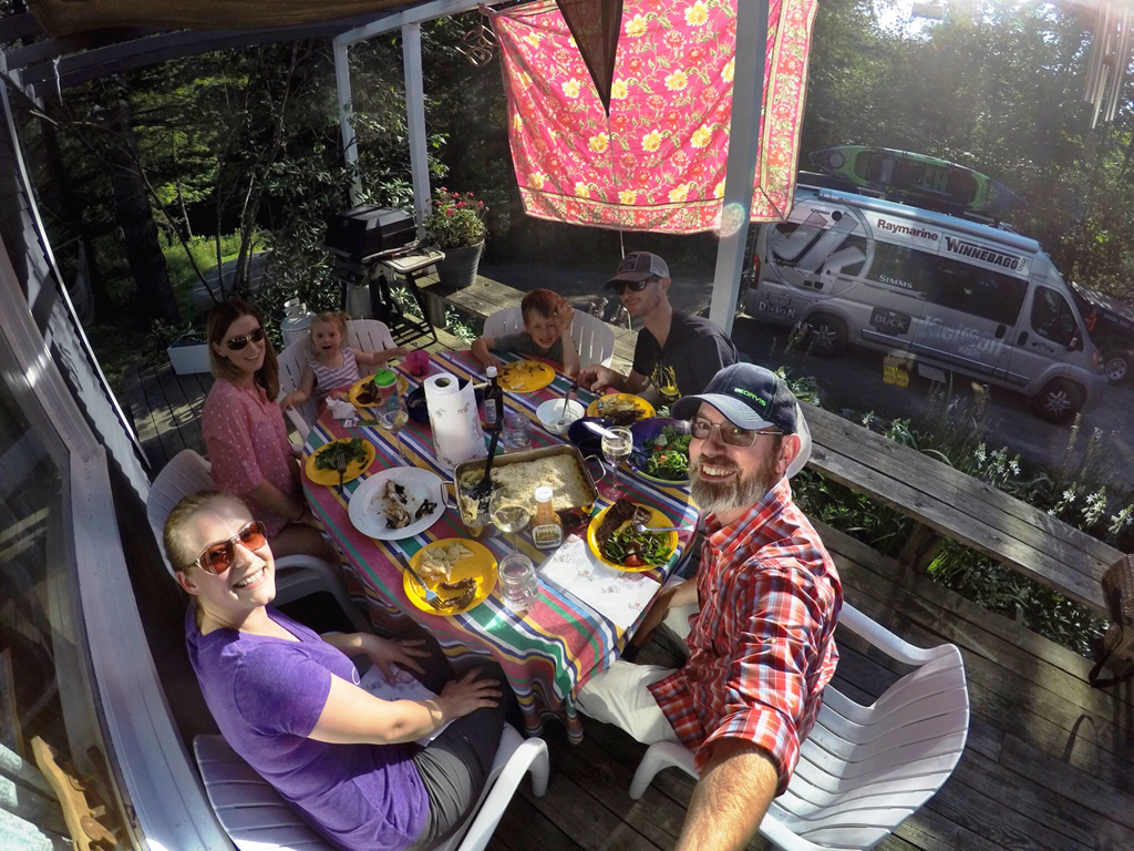 Family gathered on porch eating a meal together with Winnebago Travato visitble in the driveway.