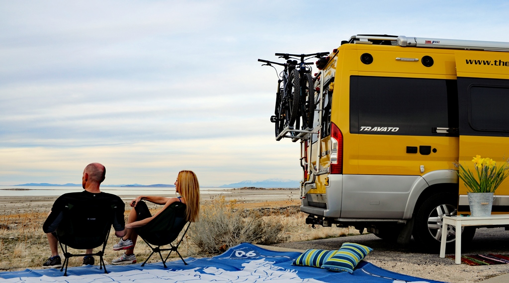 James and Stef sitting in camping chairs on the beach outside their Winnebago Travato.