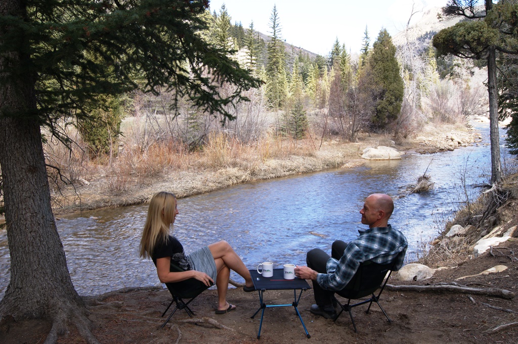 James and Stef sitting along the river in camping chairs with table between them.