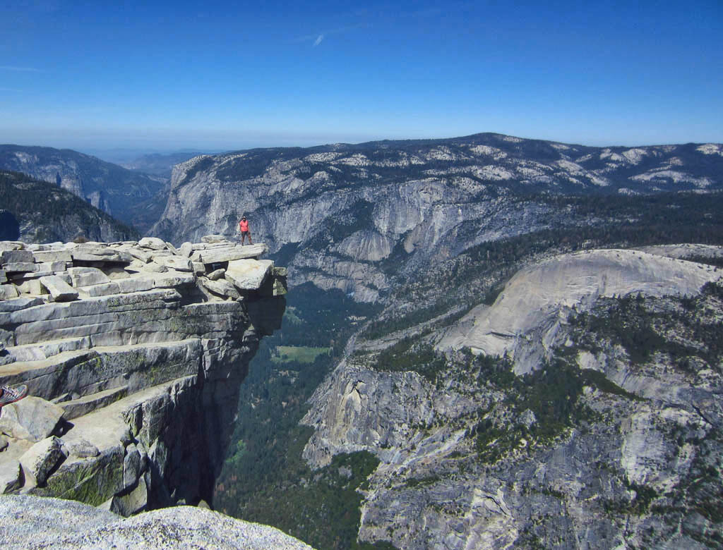 Hiker out on edge of cliff leading to tree filled valley below and mountain range surrounding.