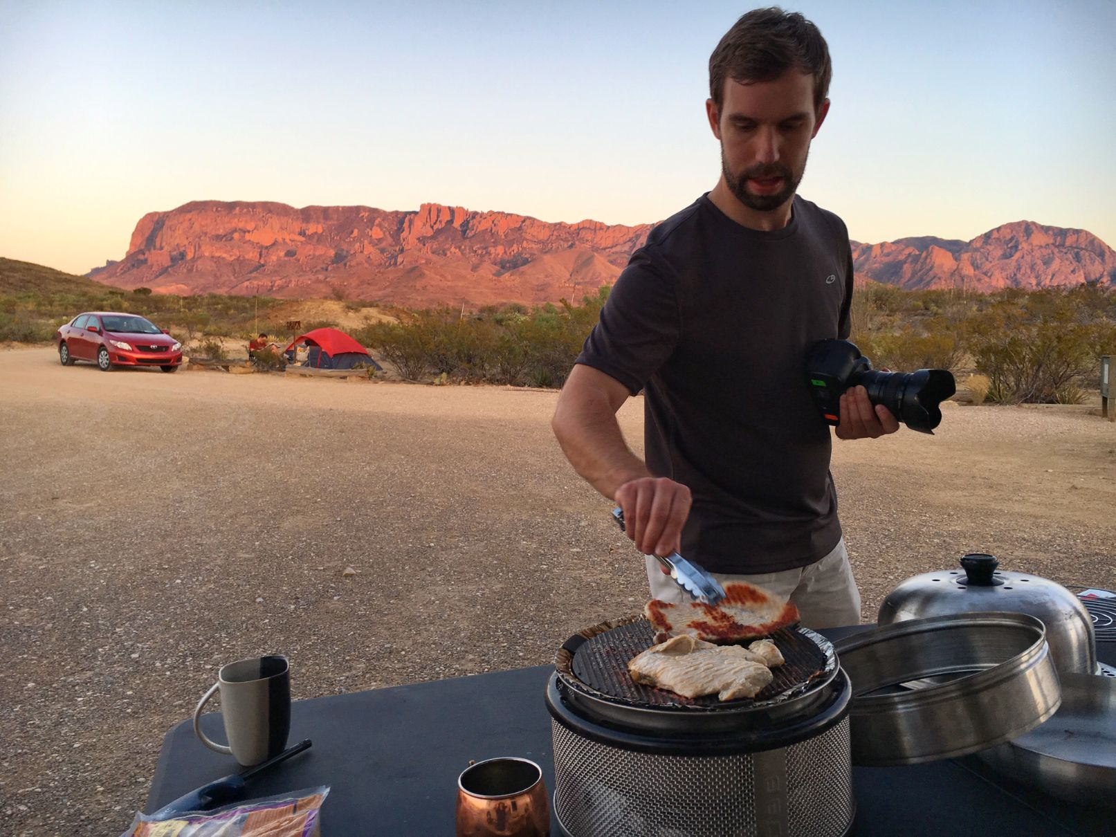 Man grilling with other campers and the mountains in the background.
