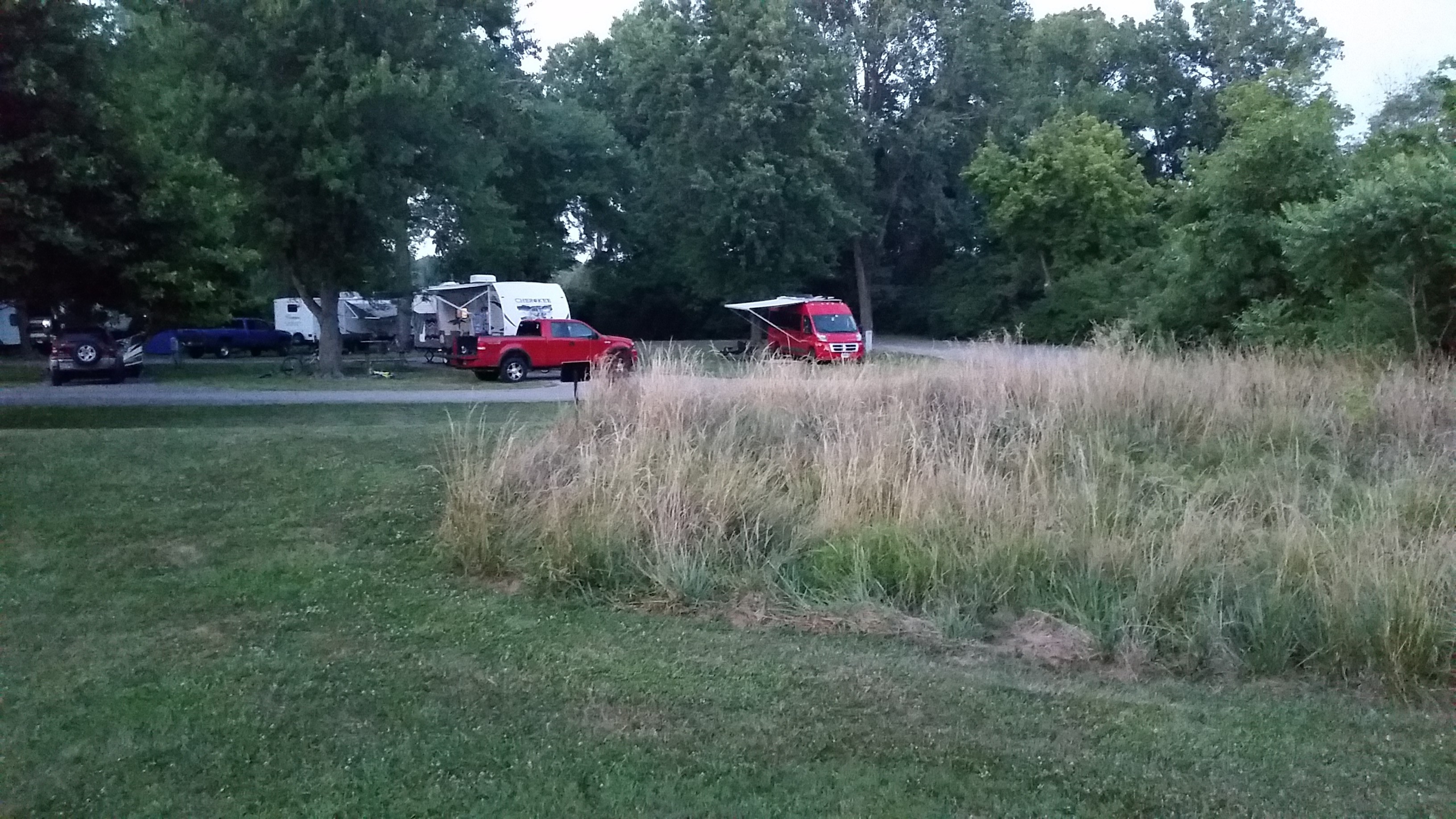 Red Winnebago Travato parked among other RVs in a tree surrounded campground.