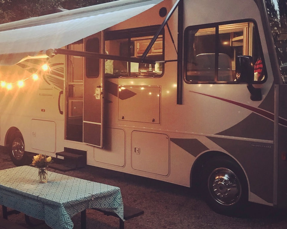 Winnebago with awning out with lights strung underneath and a picnic table with flowers on it.